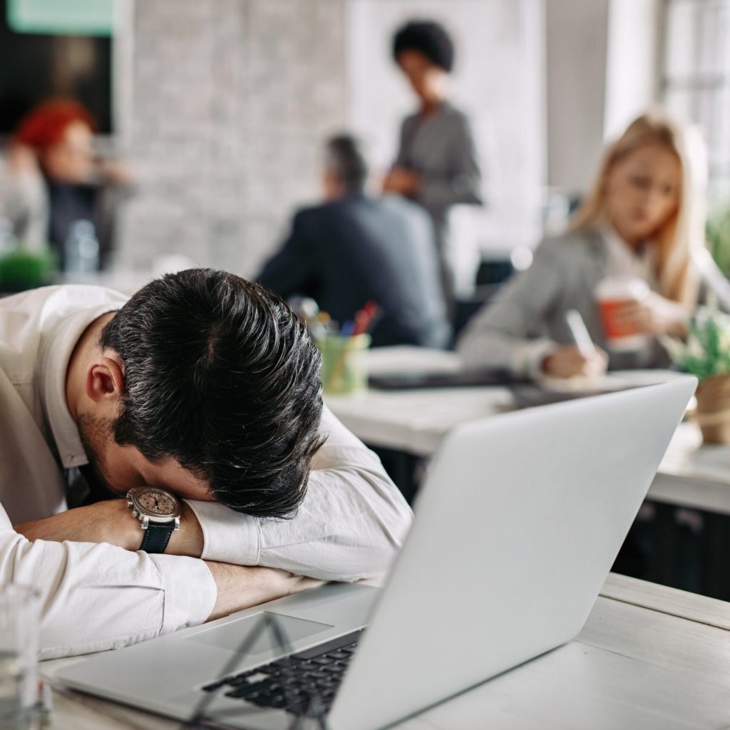 Tired entrepreneur sleeping on the table after working on computer in the office. His colleagues are working in the background.